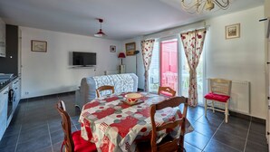 A dining area with a table set for four, a television on the wall, and a view of the outside through a window with curtains. - Les Anémones de Langrune - Avec jardin clos (Langrune-sur-Mer)