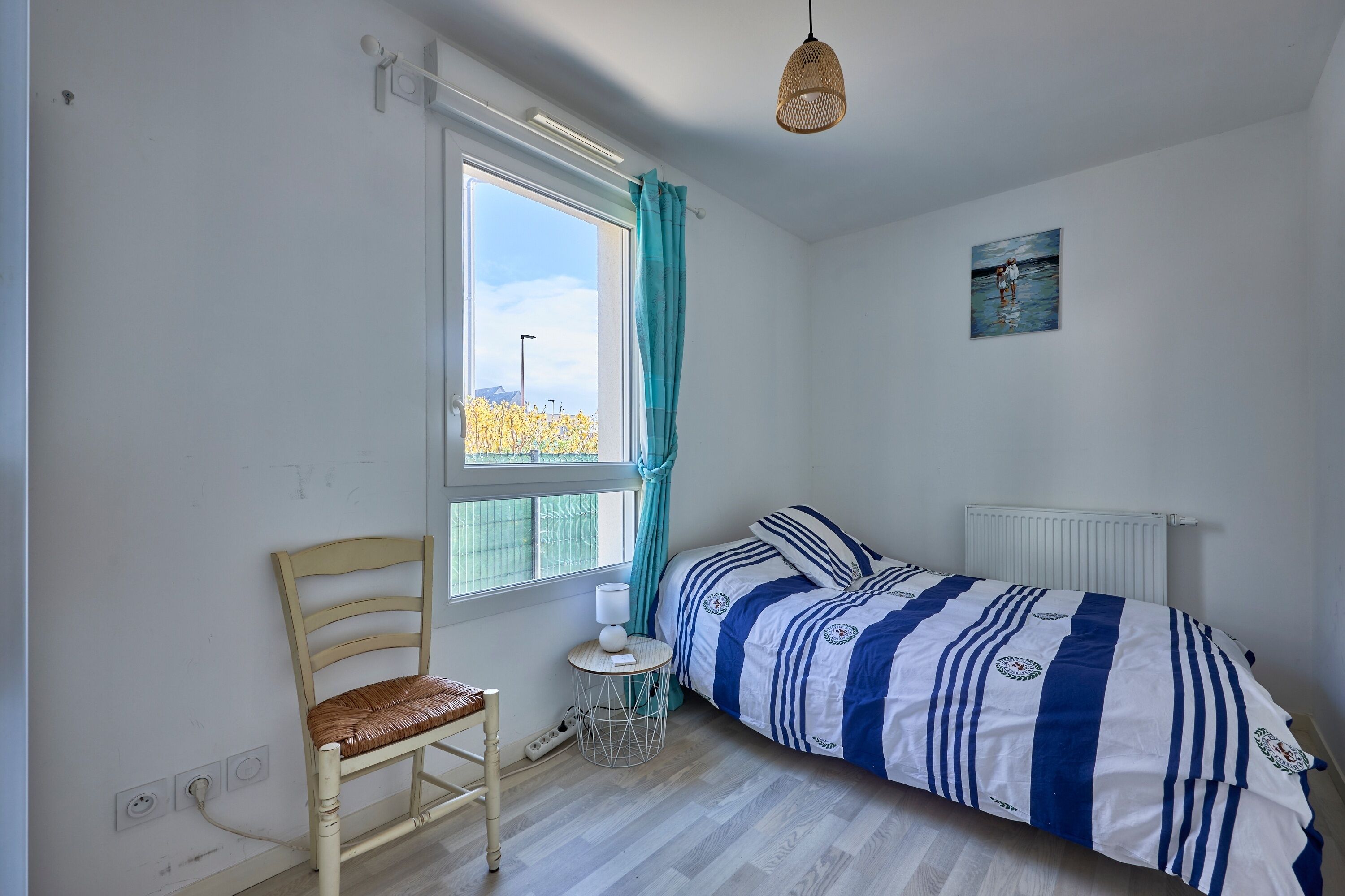 Ein Schlafzimmer mit Bett, Stuhl, einem Fenster mit Vorhängen, einem kleinen Tisch und einer Heizung.