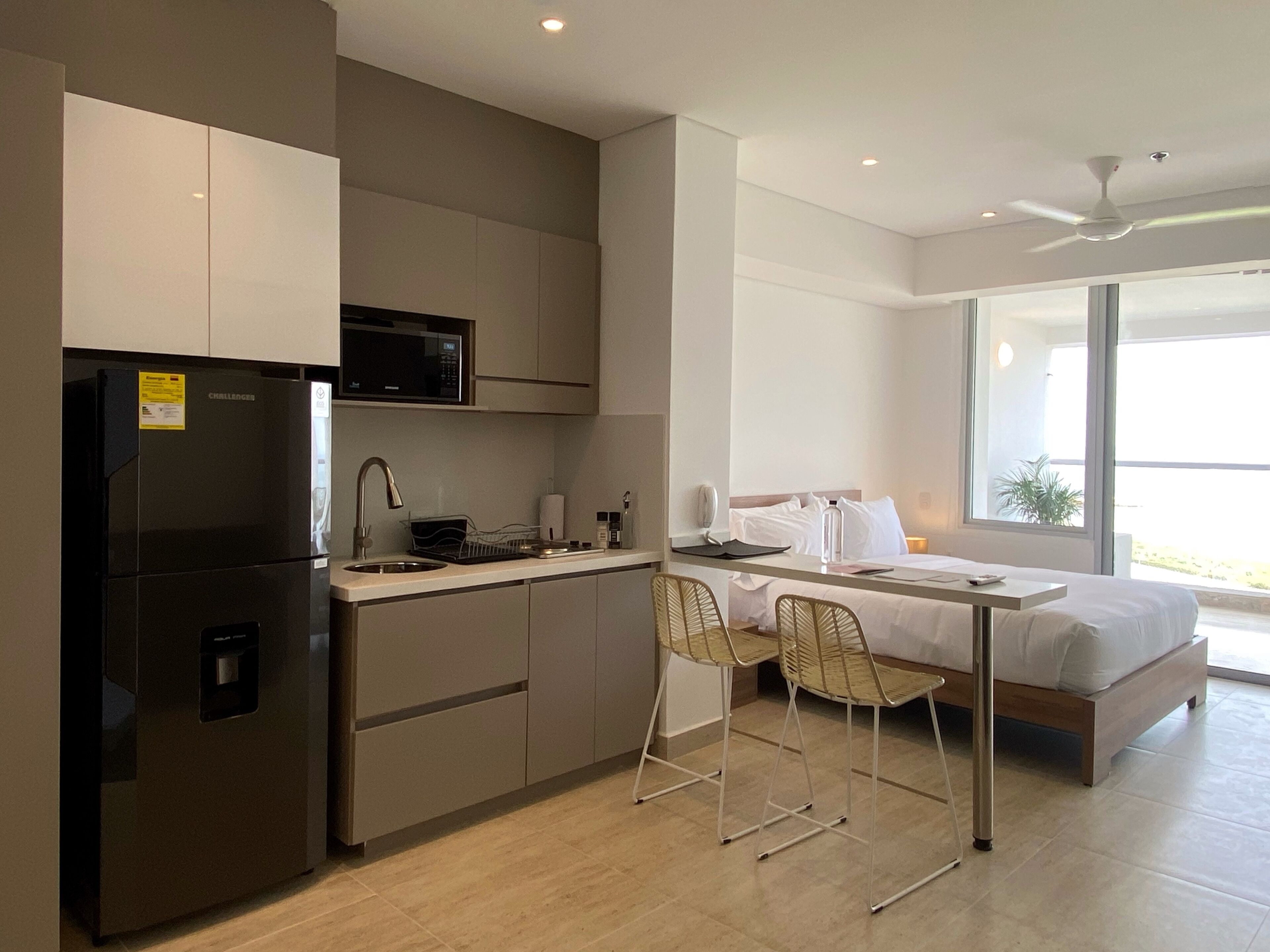 A modern kitchen with a refrigerator, microwave, and sink. There is a bed with a window view and two chairs.