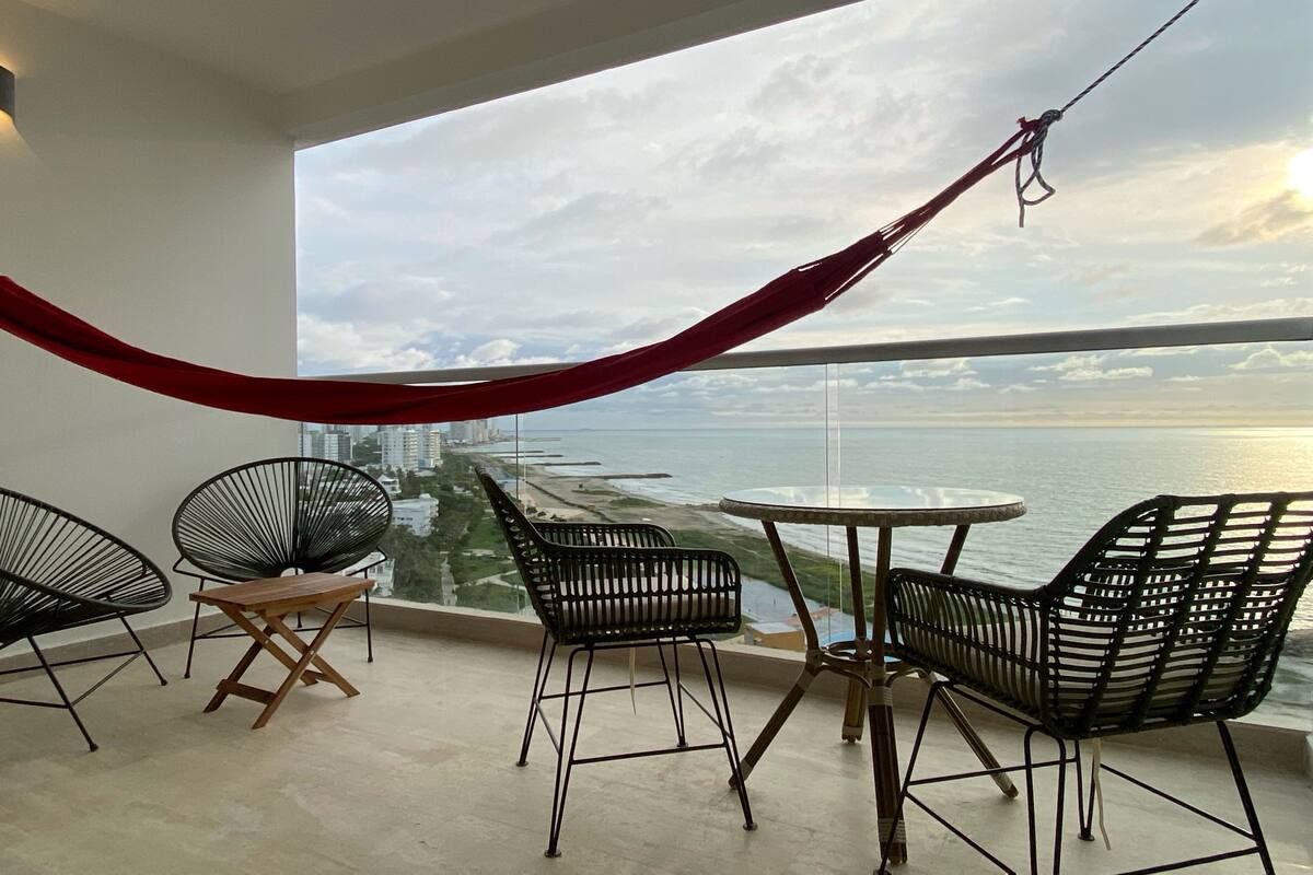 A balcony with a view of the sea, featuring a hammock, two black chairs, and a small round table.