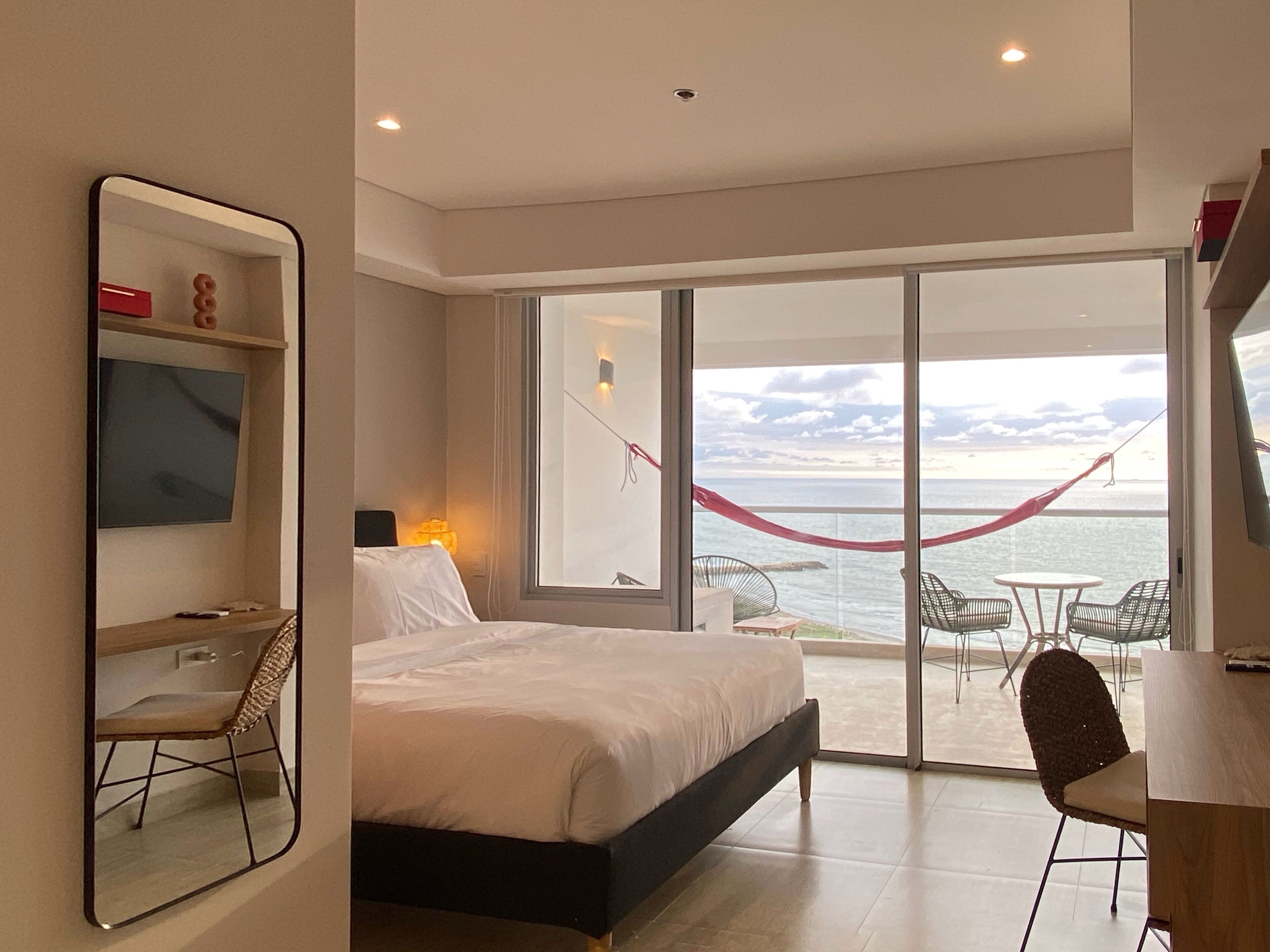 A modern hotel room with a large bed, a desk, and a balcony with a view of the sea.