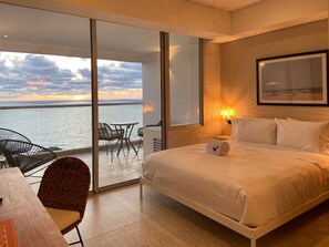 Une chambre avec un lit, un bureau et une chaise, un balcon avec une table et des chaises, et une vue sur la mer.