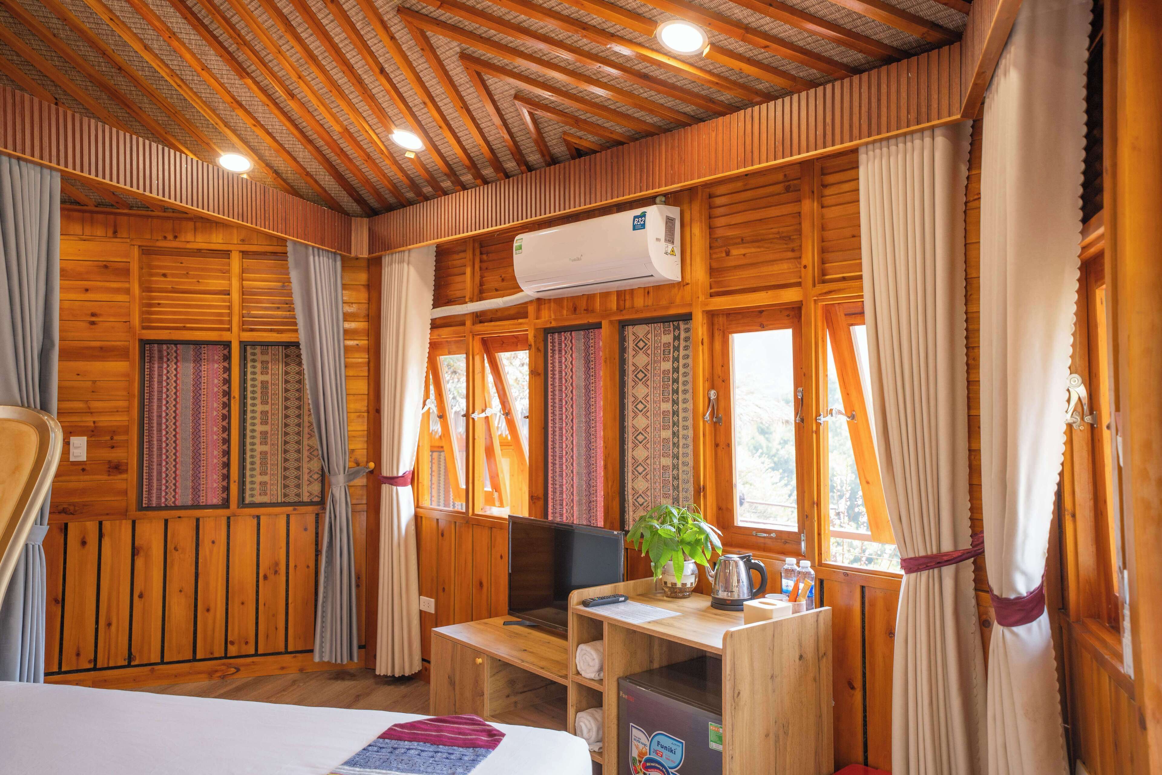 A wooden cabin room with a bed, a desk with a computer, an air conditioner, and a window with curtains.