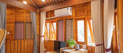 A wooden cabin room with a bed, a desk with a computer, an air conditioner, and a window with curtains.