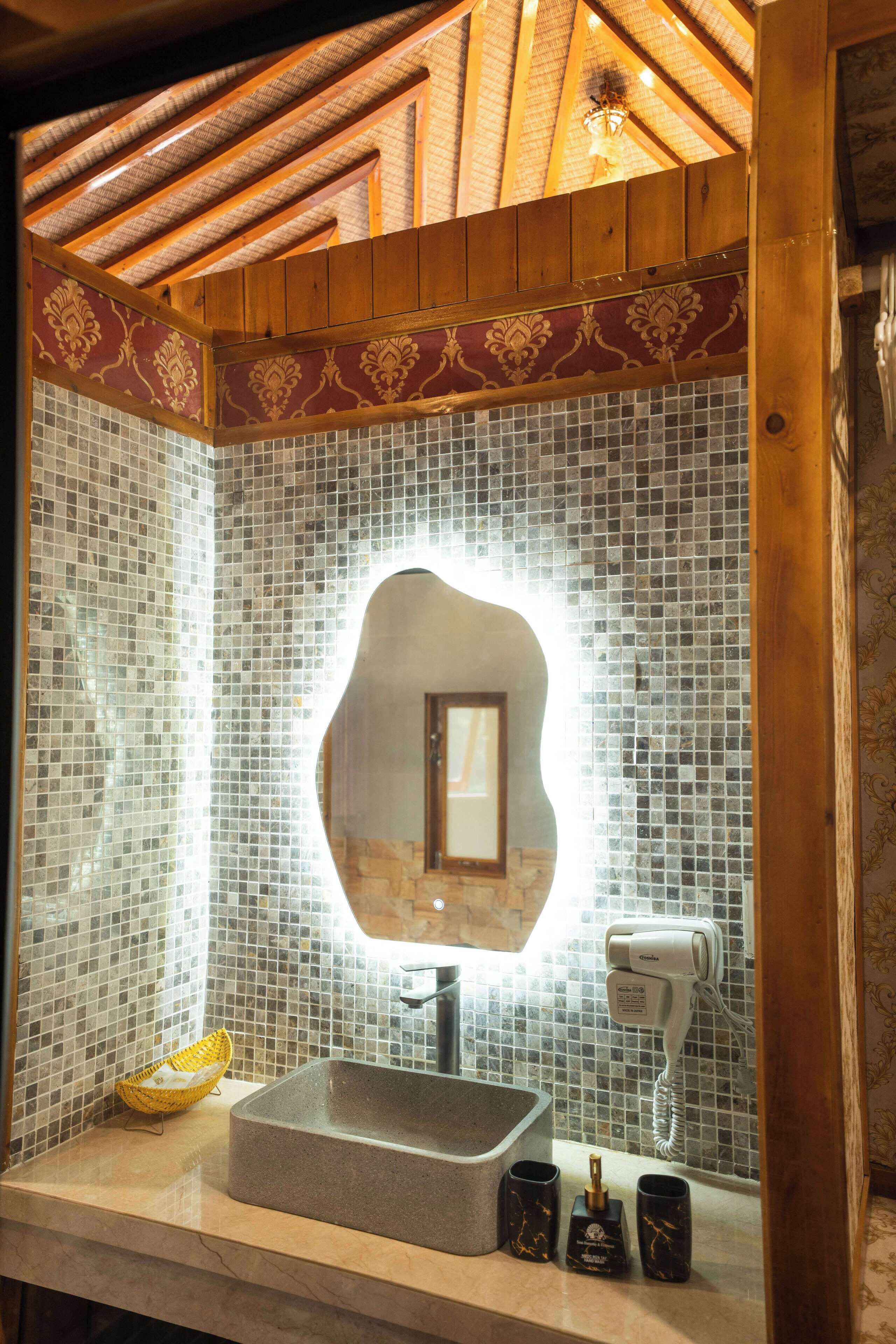 A bathroom with a tiled mosaic wall, a sink with a mirror, a hairdryer, and a yellow basket.