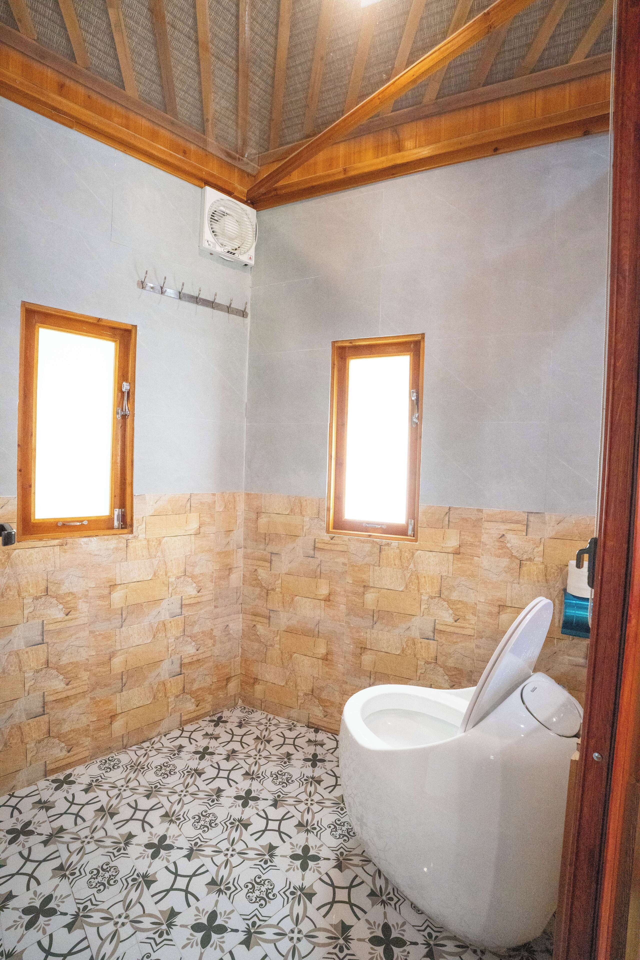 A bathroom with a stone wall, a white toilet, and two windows.