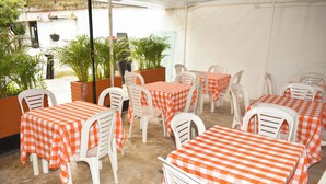 Un restaurante con mesas al aire libre cubiertas con manteles a cuadros anaranjados y blancos, sillas de plástico blanco y plantas en macetas.
