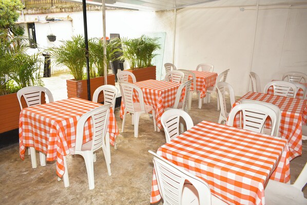 Um restaurante com mesas ao ar livre cobertas por toalhas de mesa quadriculas laranja e brancas, cadeiras de plástico branco e vasos com plantas.
