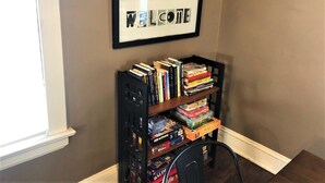 A bookshelf filled with books and board games, a framed welcome sign, and a large wall clock. - Jefferson House near the Square, Cozy & Clean, Close to Everything! (Medina)