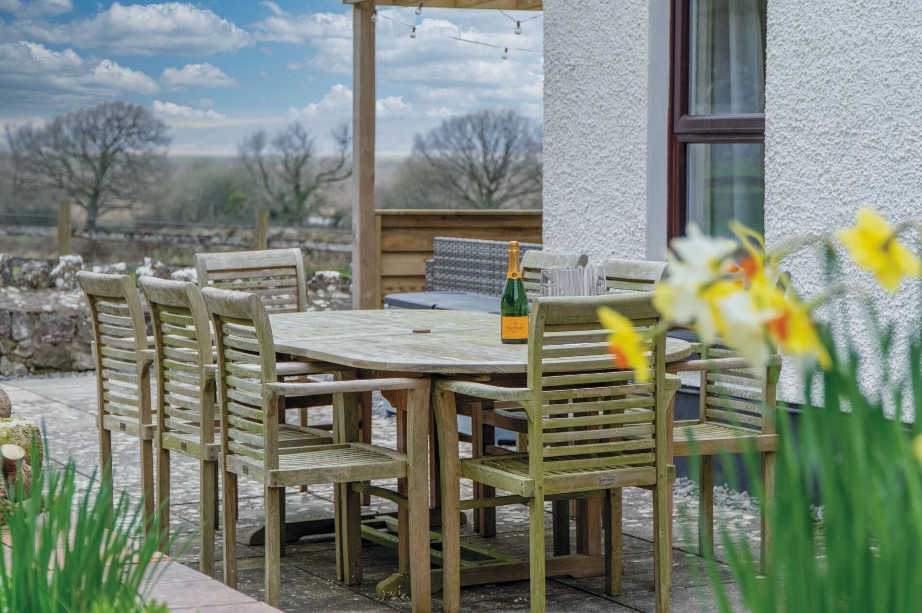 A wooden table and chairs set outdoors with a bottle of champagne, surrounded by greenery and a stone wall.