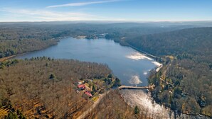 An aerial view of a lake surrounded by forested hills, a small settlement with buildings, and a bridge connecting the areas. - Domes at Catskills a lakefront glamping adventure for adults (Swan Lake)