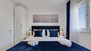 A neatly made bed with a dark blue bedspread and white pillows, two bedside lamps, and a window with blue curtains.
