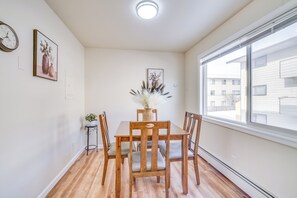 En matplats med ett träbord och stolar, en vas med torkade blommor och ett fönster med utsikt över byggnader.