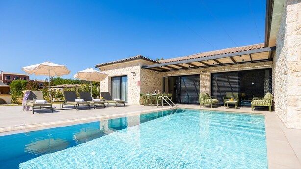 A swimming pool with lounge chairs and umbrellas, a stone building with a tiled roof, and a clear blue sky.