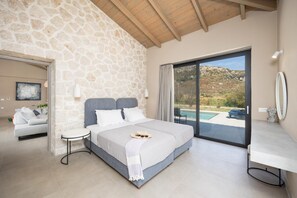 A bedroom with a stone wall, a bed with white bedding, a small bedside table, and a view of a swimming pool through a glass door.