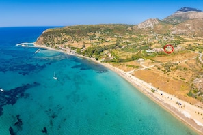 Vue aérienne d’une zone côtière avec des eaux turquoise limpides, une plage de sable et un paysage vallonné parsemé de bâtiments.