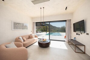 Ein modernes Wohnzimmer mit einer Couch, einem Couchtisch und einem Fernseher. Ein großes Fenster bietet Blick auf einen Pool und Hügel.