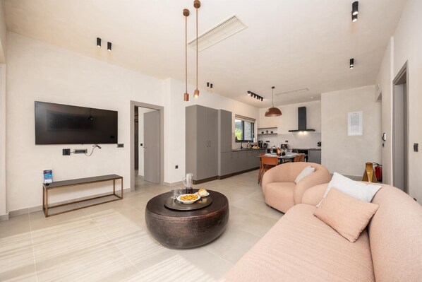 Ein modernes Wohnzimmer mit einem großen Flachbildfernseher, einem runden Couchtisch mit Snacks, einem beigen Sofa und einer Küche im Hintergrund.