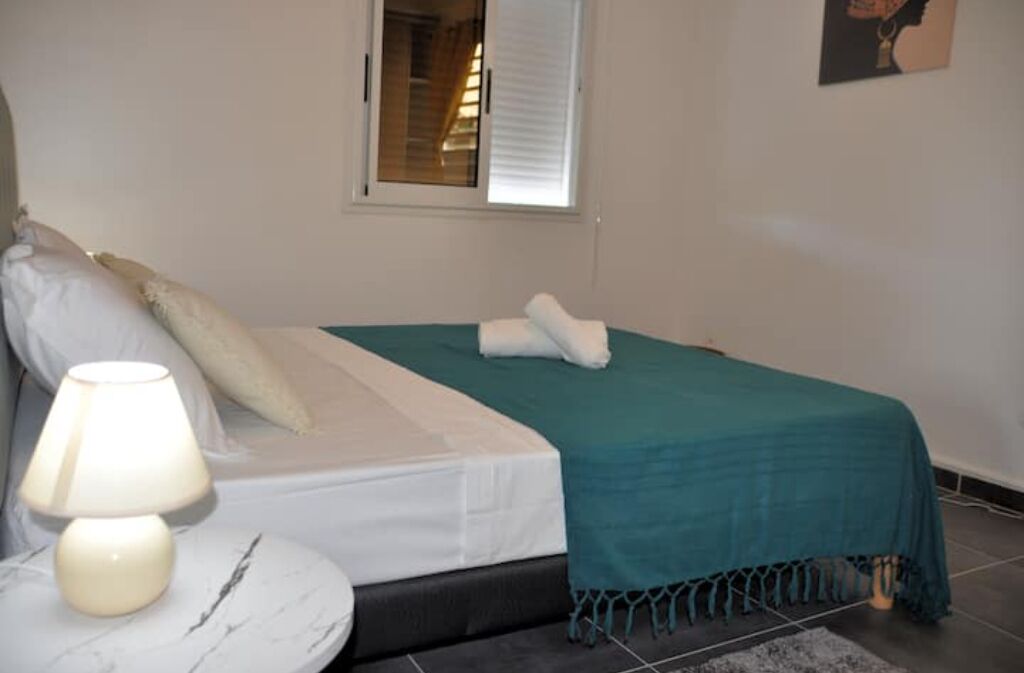 A bedroom with a bed, a nightstand with a lamp, a window with blinds, and a painting on the wall.