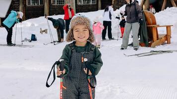 A child in a ski outfit standing in a snowy area with a wooden cabin and trees in the background.