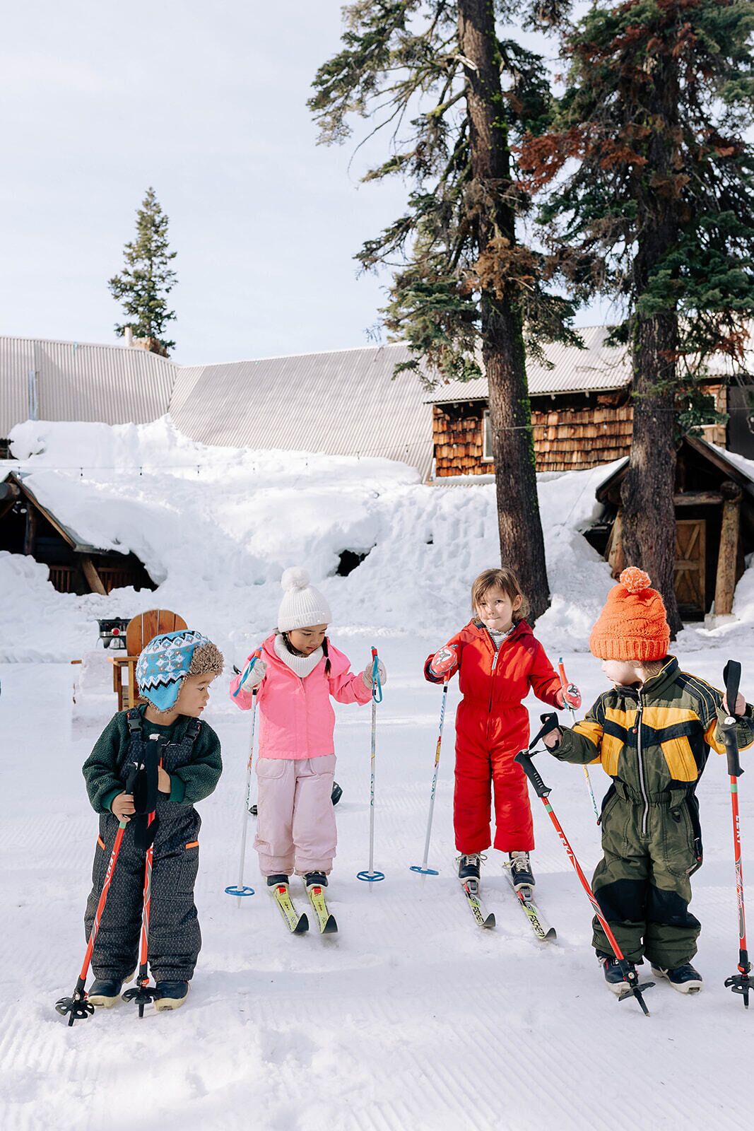Four children skiing in a snowy area with buildings in the background.