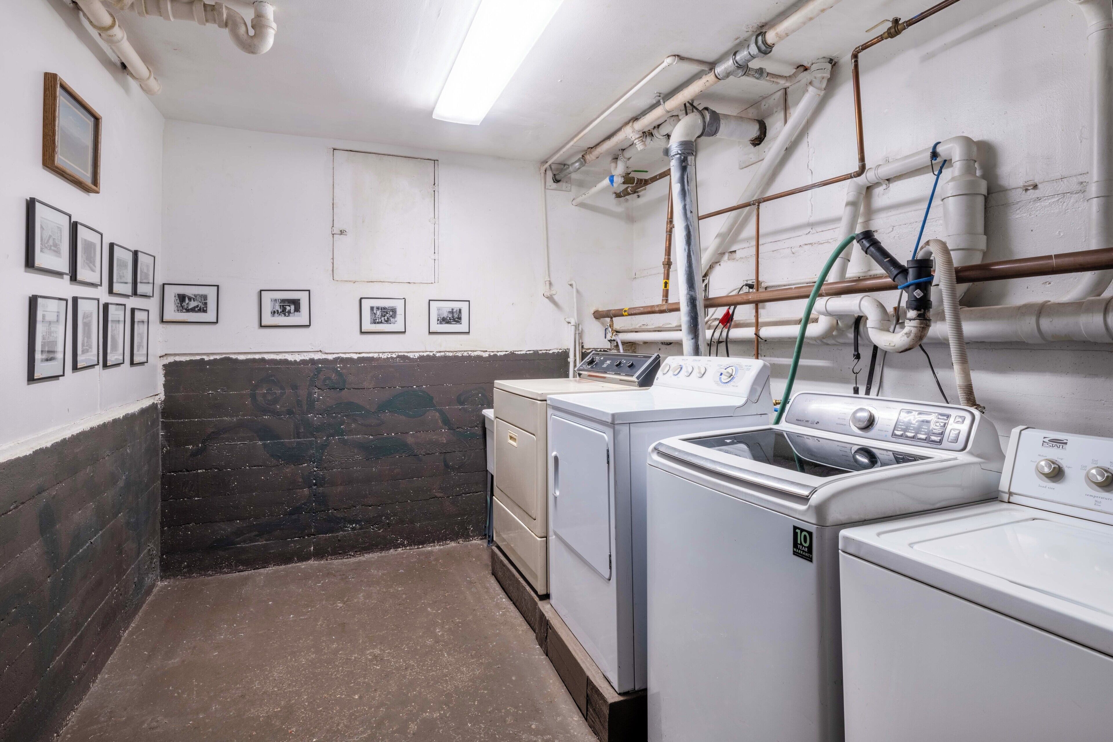 A laundry room with a washing machine, dryer, and stacked washer-dryer unit. There are pipes and a water hose on the wall, and several framed pictures on the wall.