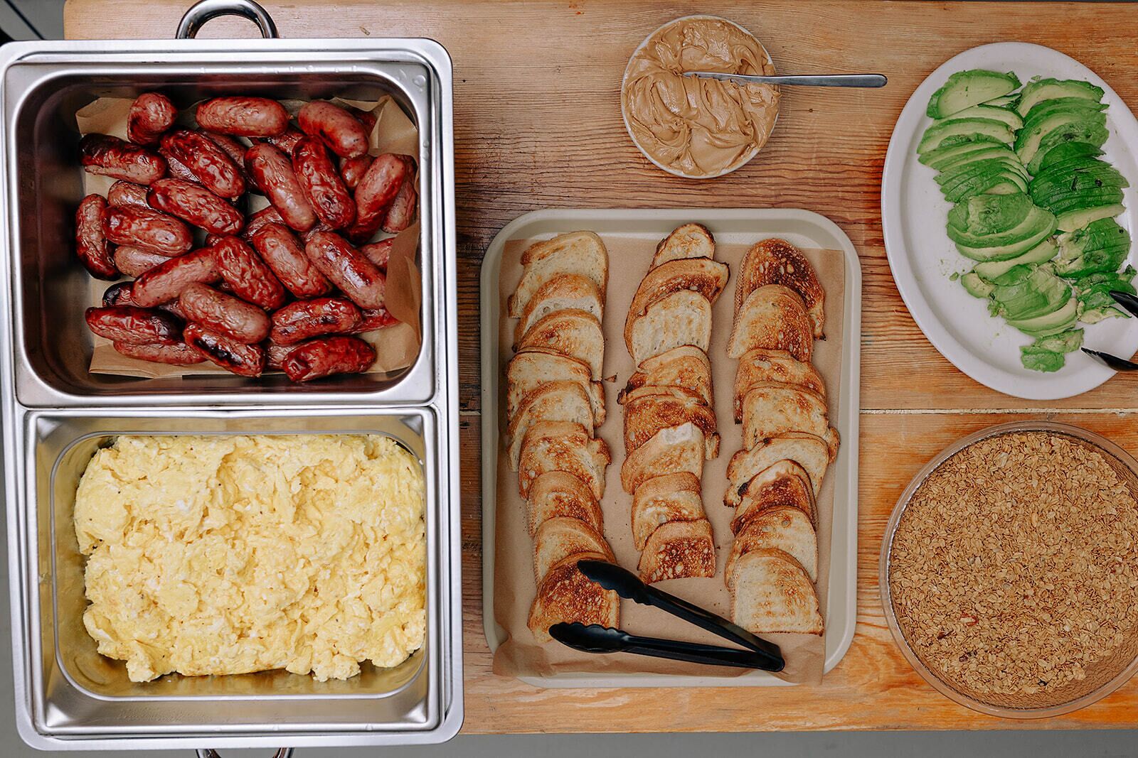 Ein Buffet mit verschiedenen Speisen, darunter Würstchen, Rührei, Toast, Avocado-Scheiben und Granola.