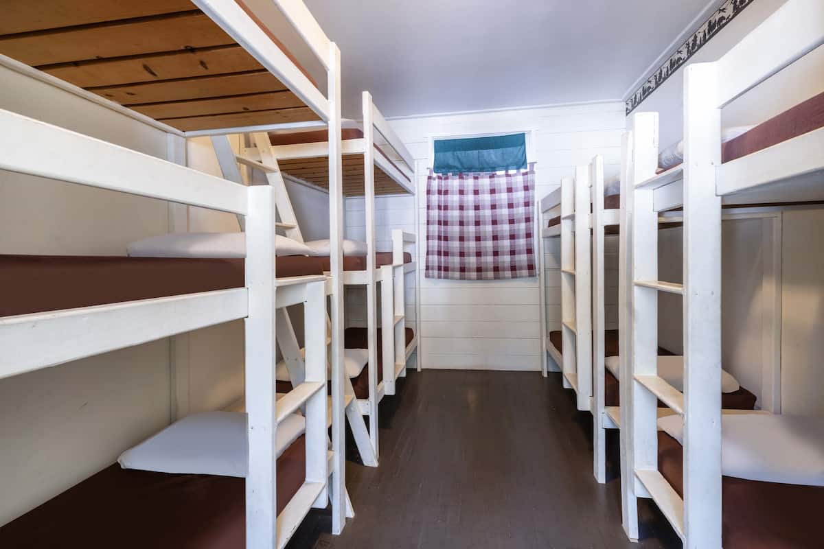 Un dormitorio con literas dispuestas en fila, una ventana con cortina a cuadros y suelo de madera.
