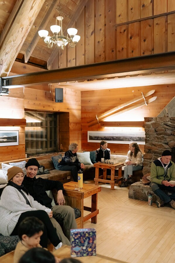 Un intérieur chaleureux en bois, avec une cheminée, une table d’échecs et un groupe de personnes assises.