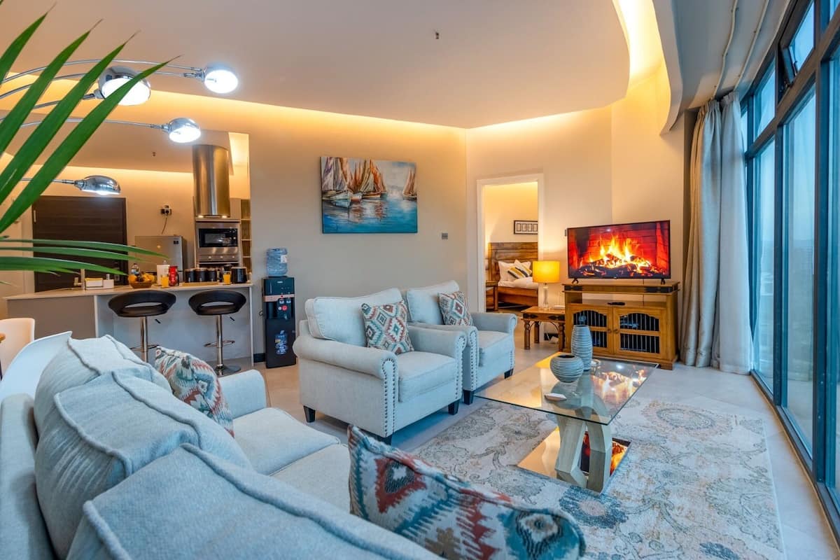 A modern living room with a sofa, armchairs, a glass coffee table, and a TV. There is a kitchen area with bar stools and a water dispenser.
