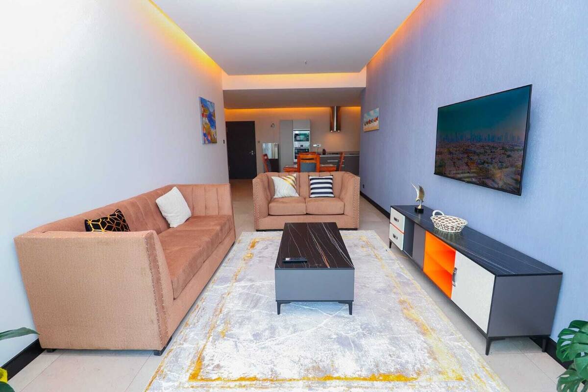 A modern living room with a sofa, a coffee table, and a TV. There is a kitchen in the background.