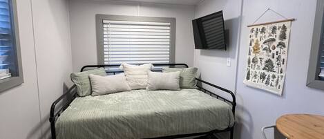 A small, well-lit bedroom with a single bed, a bedside table, a wall-mounted TV, and a window with blinds.