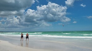 A beach with people walking, a kite surfer in the distance, and a partly cloudy sky.