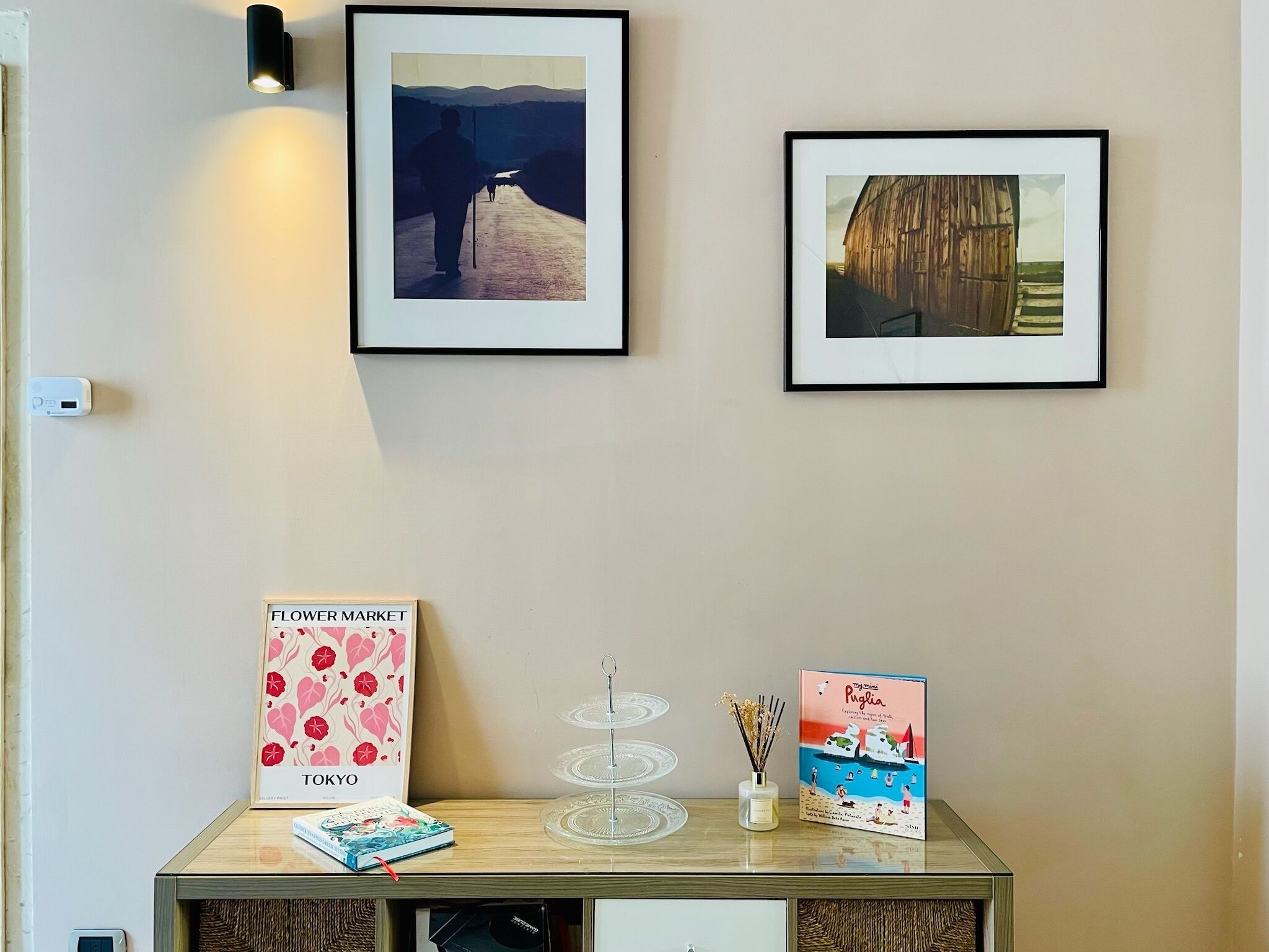 Un muro con tre quadri incorniciati, un tavolino con un libro, un vaso con fiori e una locandina incorniciata.