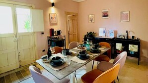 Un comedor con una larga mesa dispuesta para cuatro personas, rodeada de sillas. Hay una puerta con marco de madera, un aire acondicionado instalado en la pared y obras de arte enmarcadas.