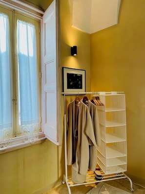 Una habitación con ventana, un perchero con ropa colgada, un cuadro en la pared y un mueble de almacenamiento blanco.