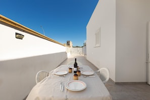 Et spisebord dækket til fire med vin, tallerkener, bestik og træskåle på en terrasse med klar himmel.