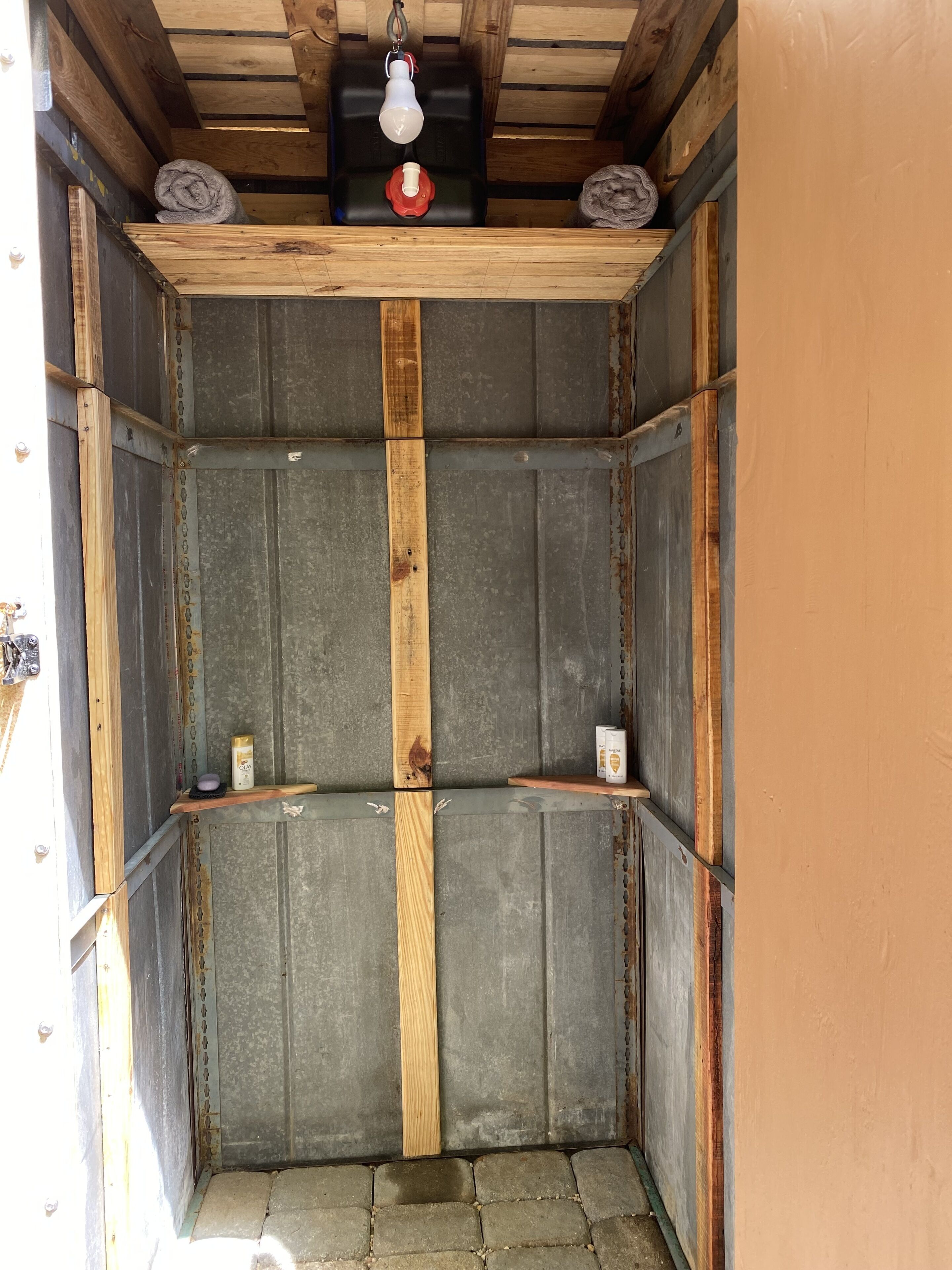 A compact, enclosed space with a wooden ceiling, metal walls, and a small shelf with a light fixture and a red object.