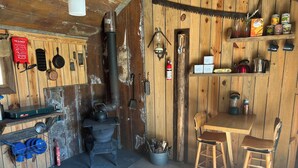 Een rustiek houten chaletinterieur met een houtkachel, een branddeken, kookgerei en een klein eethoekje met tafel en stoelen.