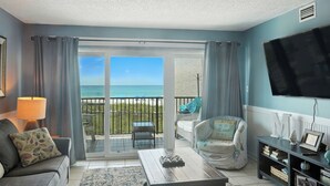 Ein Wohnzimmer mit Blick auf den Strand, eine Couch, ein Couchtisch, ein Fernseher und ein Bücherregal.