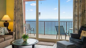 Habitación con balcón que da al mar, amueblada con sofá, sillas y una mesa con un jarrón de flores.