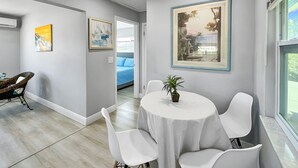 Ein modernes Zimmer mit einem runden Tisch, weißen Stühlen und einer Zimmerpflanze. An den Wänden befinden sich gerahmte Kunstwerke und ein Fenster mit Aussicht.