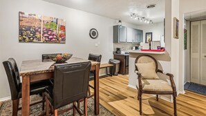 A dining area with a wooden table, chairs, and a decorative bowl. A kitchen is visible in the background with cabinets and a countertop.