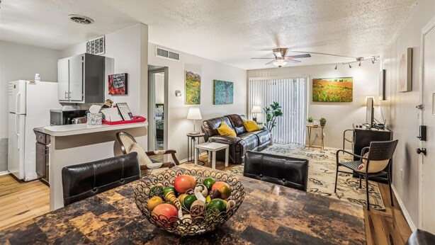 A modern living room with a dining area, featuring a kitchen with white cabinets and a refrigerator. There is a ceiling fan, a dining table with a fruit bowl, and a sofa with cushions.