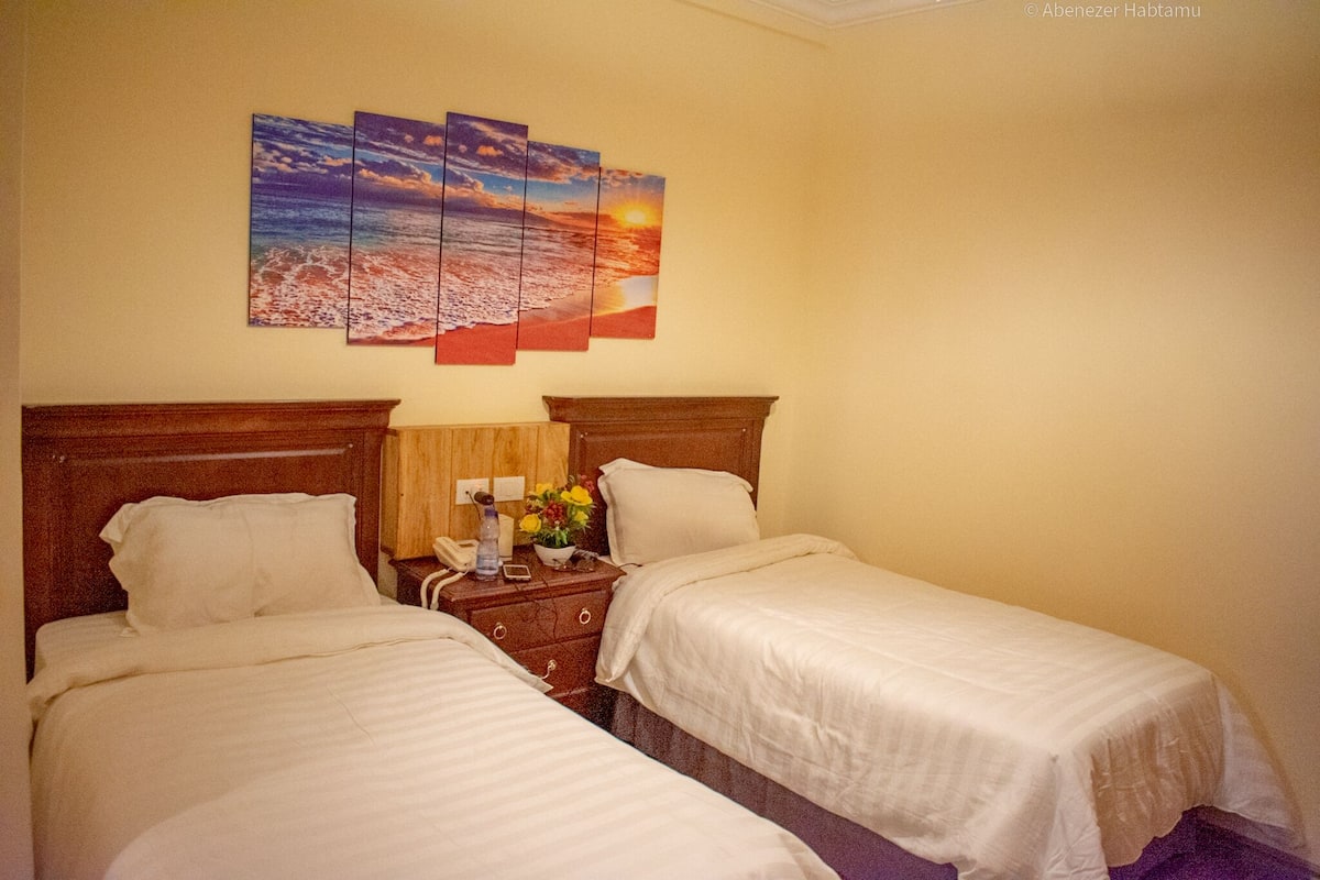 A hotel room with two beds, a wooden headboard, a nightstand with a lamp and flowers, and a sunset painting on the wall.