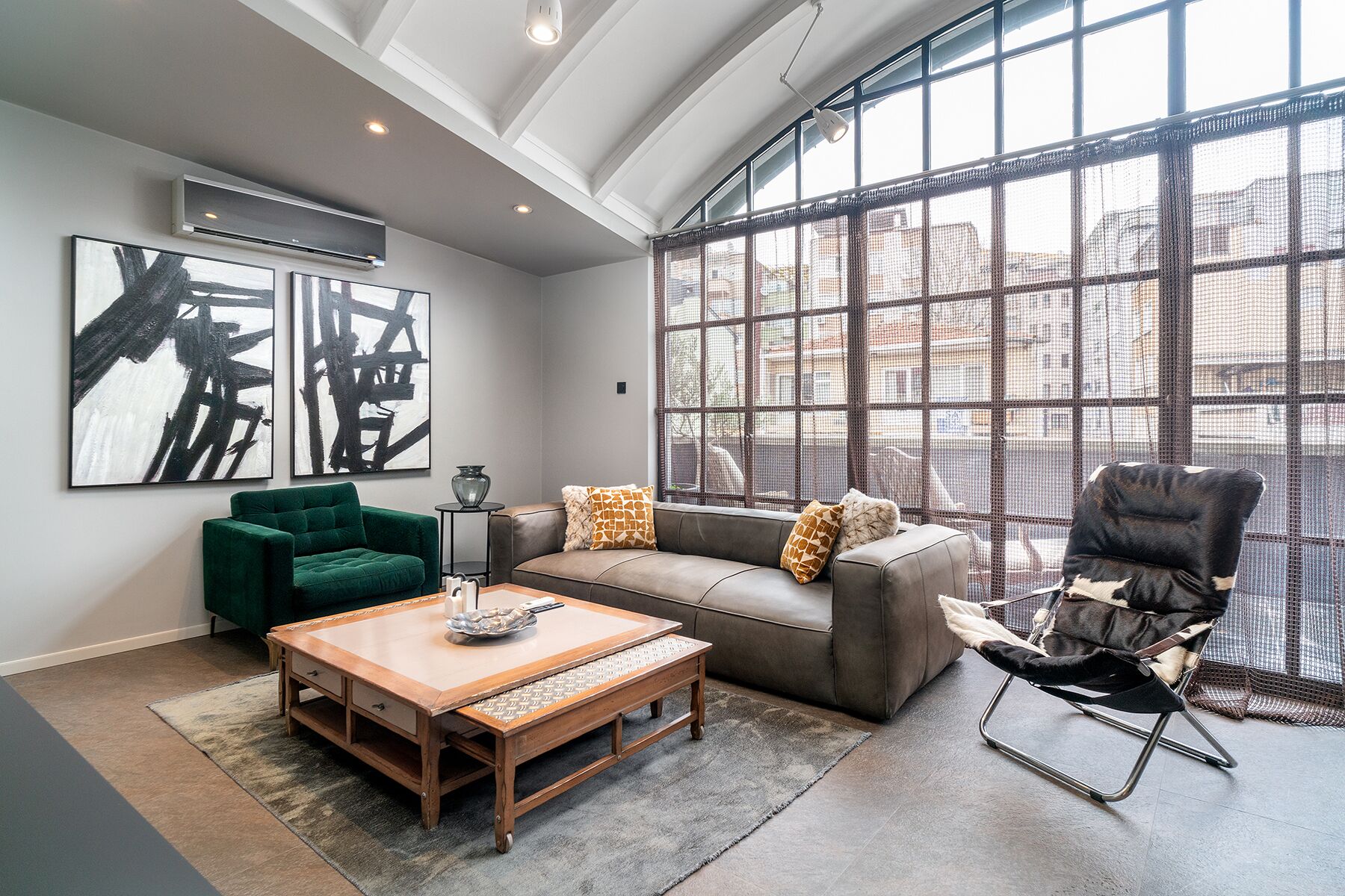 A modern living room with a green sofa, a brown leather couch, and a black chair. There is a wooden coffee table and a large window with a view of the city.