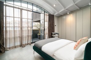 Une chambre moderne dotée d’une porte en verre incurvée donnant sur un balcon, d’un lit avec une tête de lit recouverte d’un tissu vert et d’une petite table accompagnée d’une chaise.