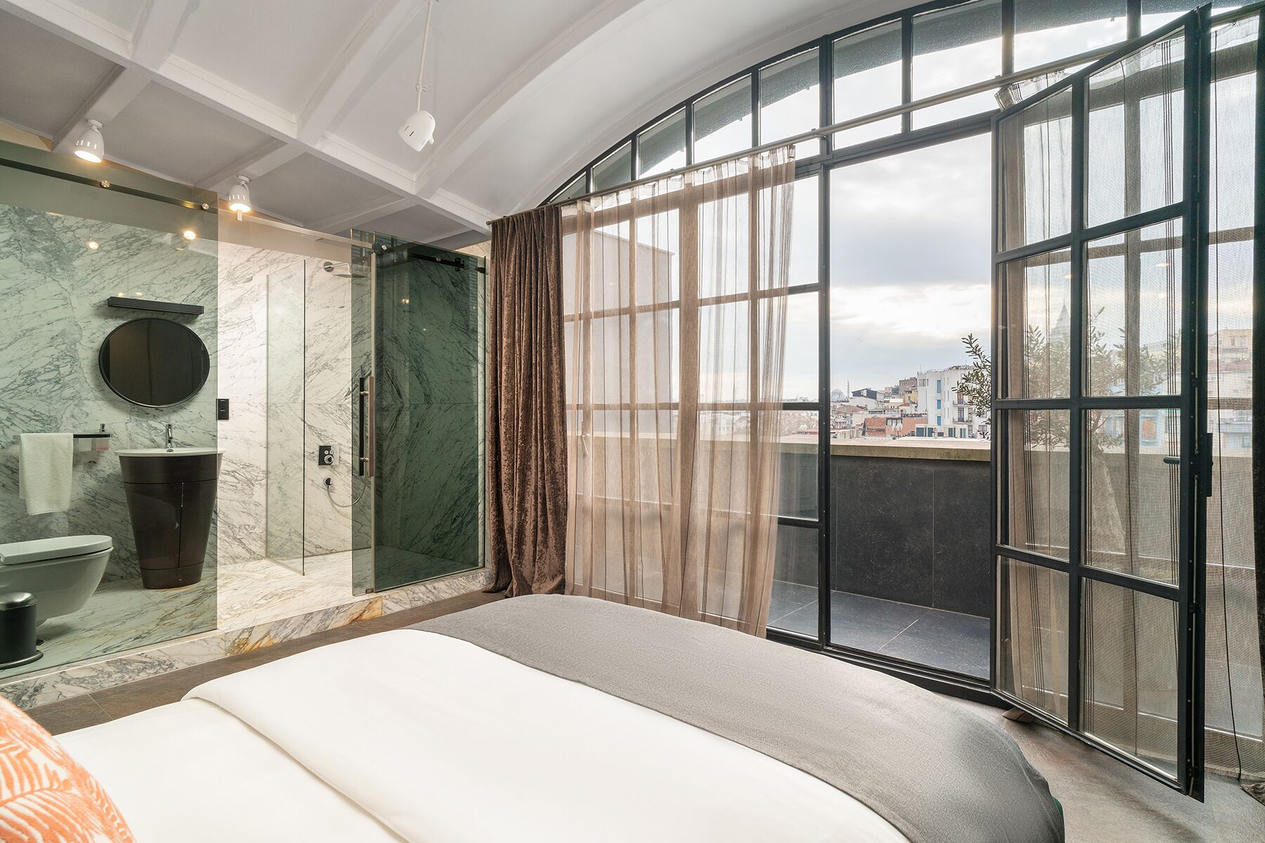 A modern bedroom with a large bed, a glass shower, and a view of the city through large windows.