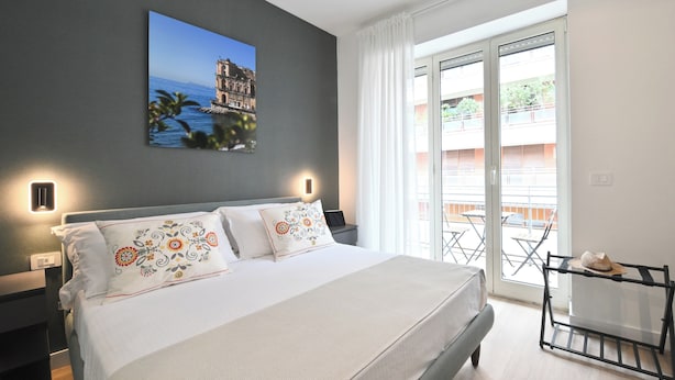 A bedroom with a bed, a nightstand, a wall-mounted lamp, a balcony with a table and chairs, and a view of a building and water. - Ponte di Tappia Suites (Naples)