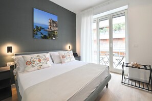 A bedroom with a bed, a nightstand, a wall-mounted lamp, a balcony with a table and chairs, and a view of a building and water. - Ponte di Tappia Suites (Naples)